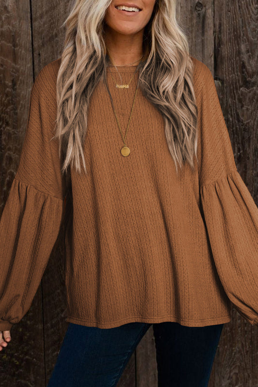 Woman wearing a brown textured long-sleeve blouse with bell sleeves, blue jeans, and a necklace with a round pendant against a wooden background.