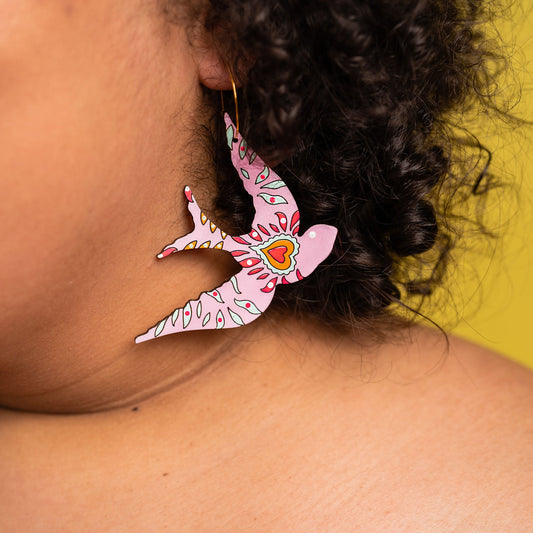 Colorful bird-shaped earring with intricate pink, red, and orange patterns worn by a person with curly hair, against a yellow background.