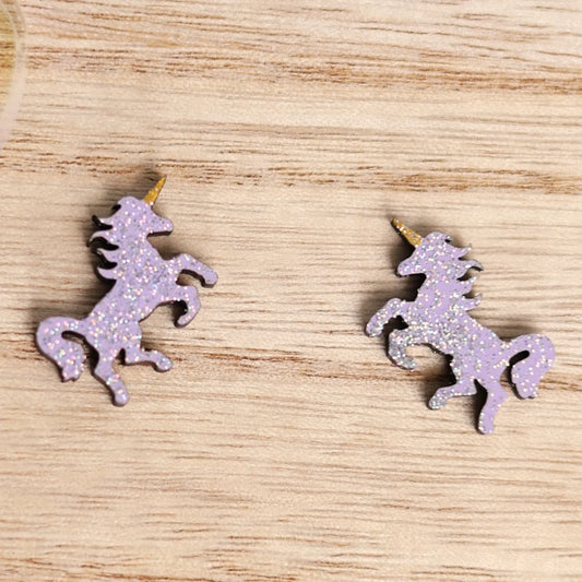 Purple unicorn stud earrings with glitter accents displayed on a wooden surface.
