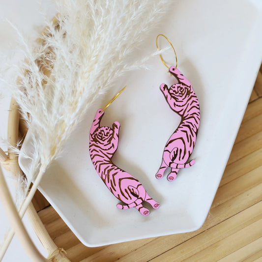 Pink tiger-shaped earrings with gold hooks displayed on a white dish, alongside decorative pampas grass on a wooden surface.