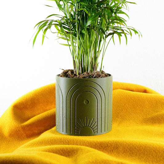 Boho-style green planter with sun and arch design, holding a leafy green plant, placed on a bright yellow cloth.