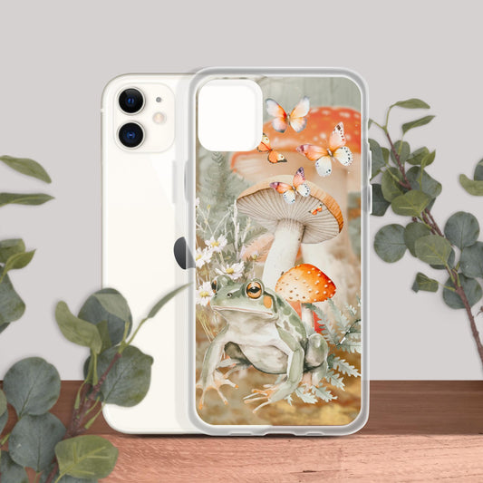 Clear iPhone 11 case featuring a design with a frog, mushrooms, daisies, and butterflies, shown with an iPhone on a wooden surface with green leaves in the background.