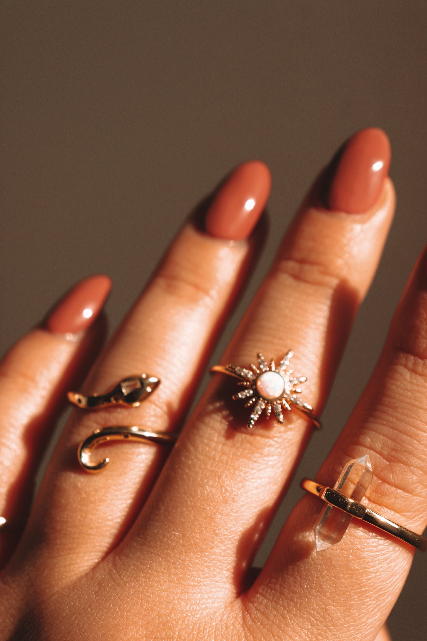 Gold rings on fingers with oval nails painted brown. Rings include a snake design, a starburst with a white gem, and a crystal point.